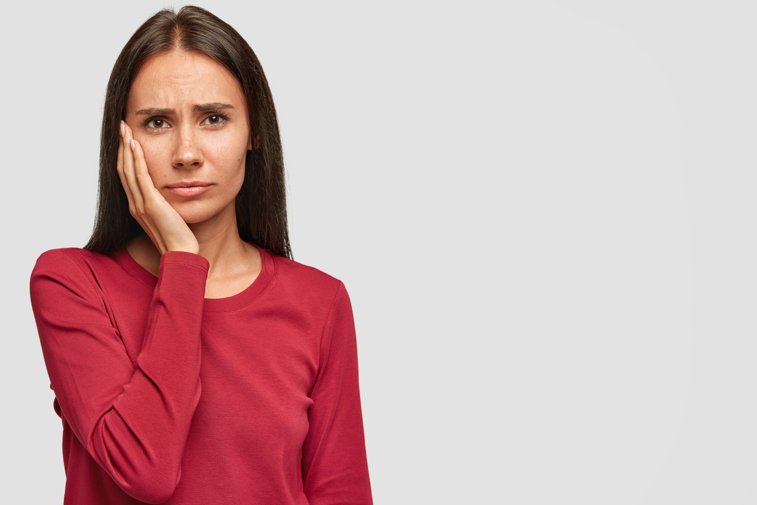 Indoor shot of sad unhappy European woman with unhappy expression, keeps palm on cheek, wears casual red sweatshirt, isolated over white background with copy space for your promotional content